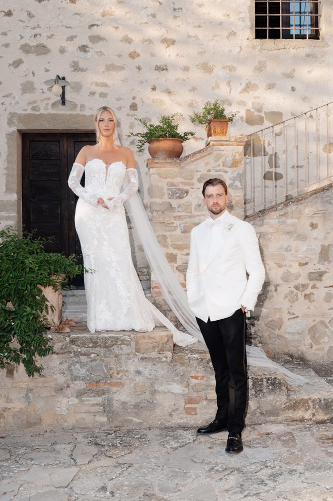 Real Bride Maartje & Rik – bruid en bruidegom poseren stijlvol bij een historisch stenen gebouw; Maartje draagt een elegante fitted trouwjurk met verfijnd kant, off-shoulder mouwen en lange sluier, Rik in een klassiek wit colbert voor een tijdloze en luxe uitstraling.